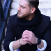 Man with a dark coat and beard sits with hands clasped, looking to the left, in a thoughtful pose.