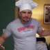 Man wearing a white chef hat and light gray T-shirt with red lettering, standing indoors with furniture in the background.