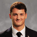 Professional headshot of a young man in a dark suit and tie, against a neutral gray background.