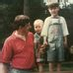 Two adults and a small child standing on a wooden play structure in a park, with trees in the background.